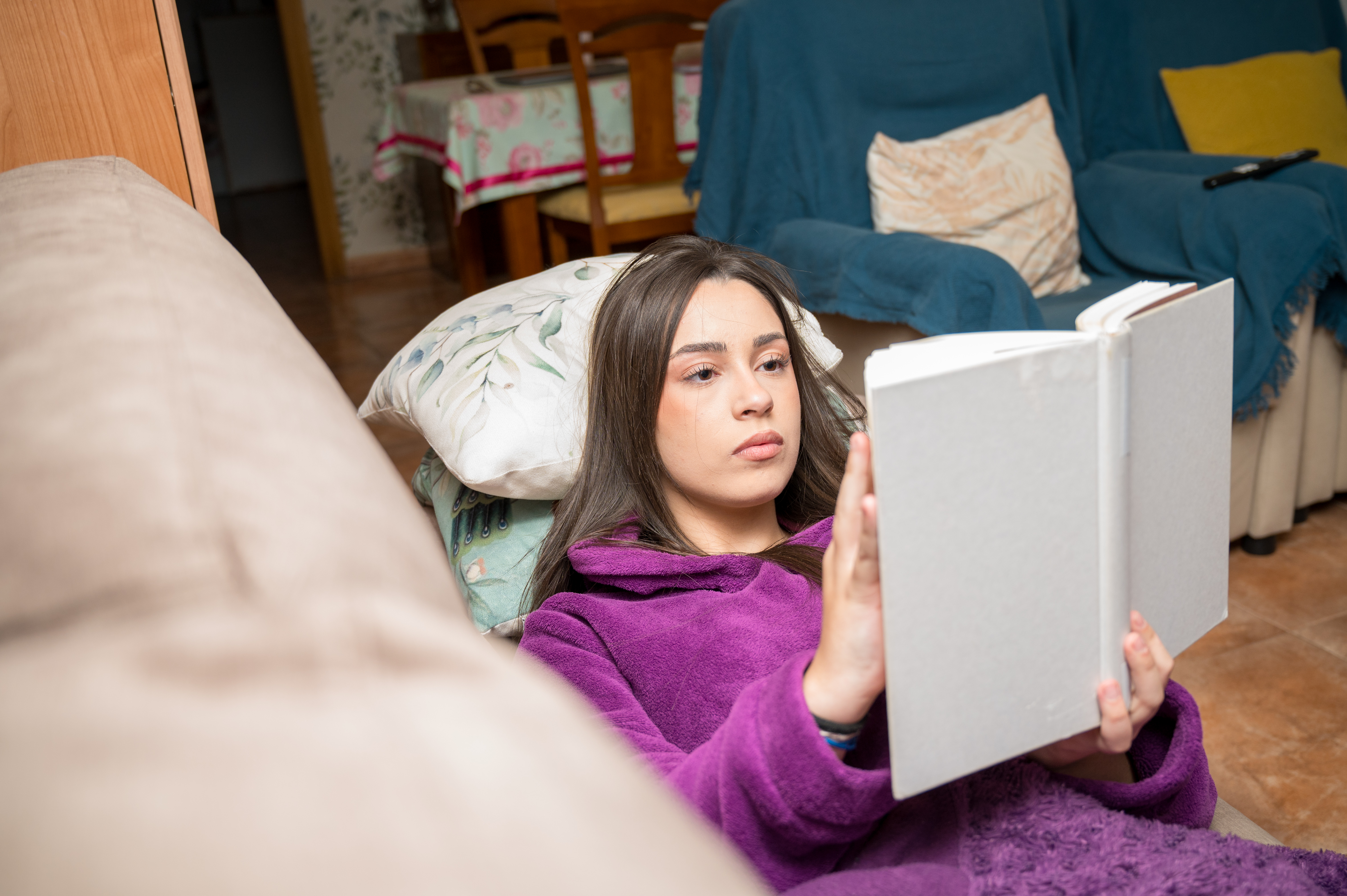 Teenage girl lying on a sofa reading a book with a focused expression in a cozy domestic living space
